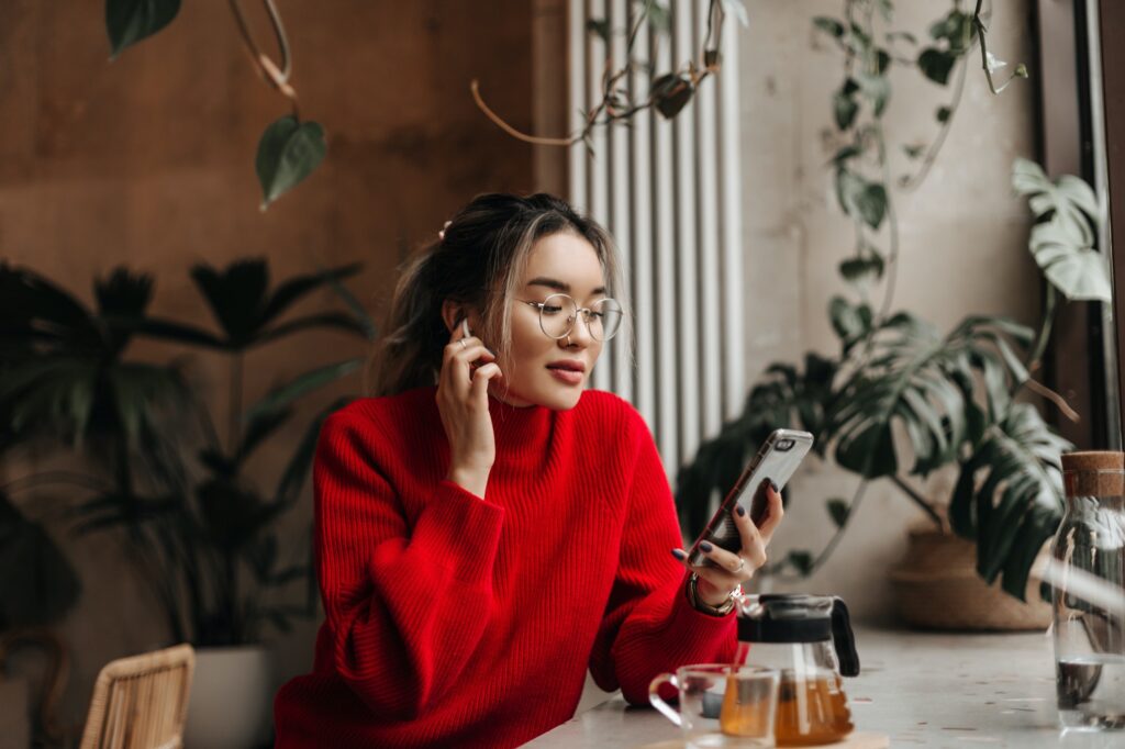 Girl in red oversized outfit and glasses holds smartphone. Asian ordered tea and sits in cafe on ba